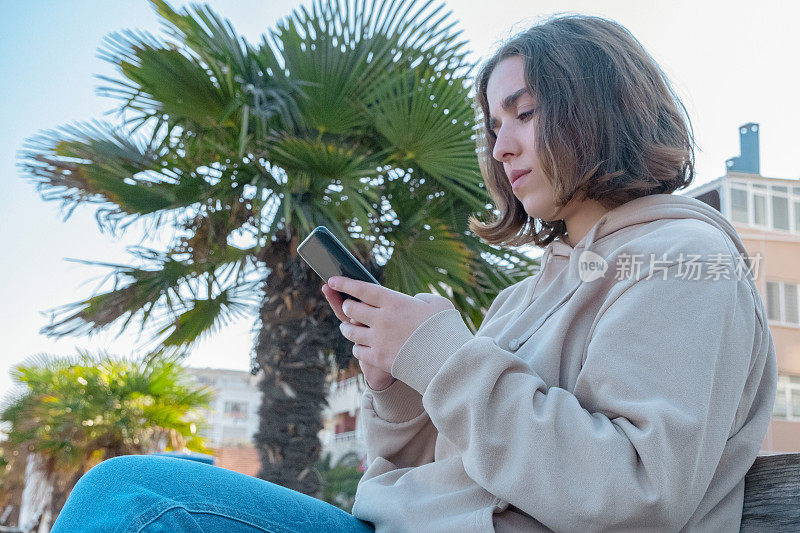 一名年轻女性坐在长凳上用手机发短信的特写。她附近有一棵棕榈树。