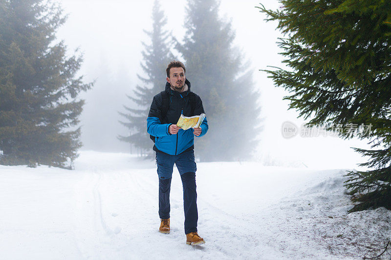 年轻的男性徒步旅行者穿着蓝色的夹克，在雾蒙蒙的雪山上走着，手里拿着地图