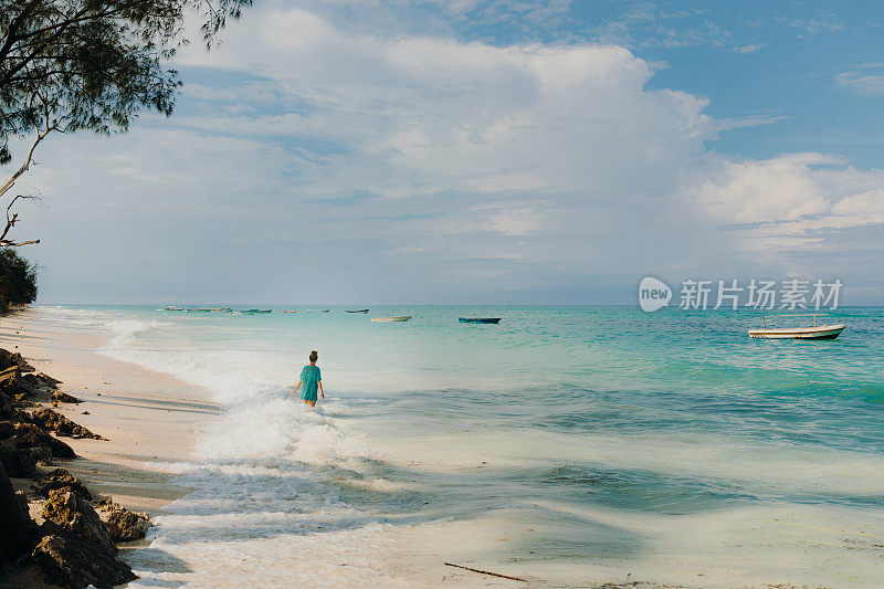 在桑给巴尔岛，一名女性游客在碧绿的海水和小船的沙滩上观赏日落