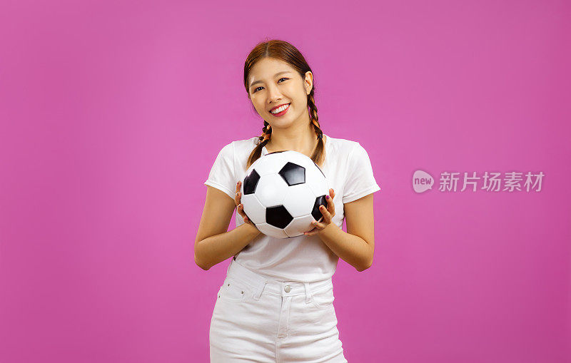 年轻的亚洲女子孤立的粉红色背景微笑着看着镜头，手里拿着一个足球，国际足联世界杯概念