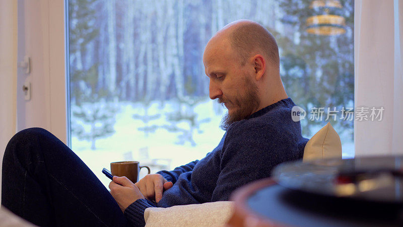 一个留着胡子的野蛮男人坐在家里的沙发上，通过智能手机上网，喝咖啡。中年男人的孤独。在家里过着单身隐士的简单生活。
