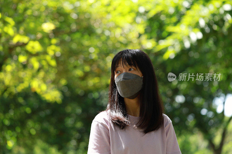 年轻的亚洲女子戴着防护口罩站在城市公园，周围都是绿色植物，她望向远方，满怀希望。