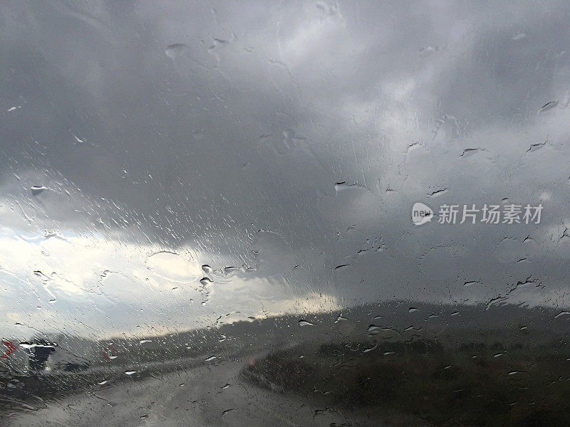 下雨天开车。雨点落在汽车挡风玻璃上，多云的天空和空旷的道路背景