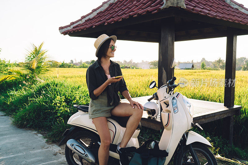 有趣的女旅行者手里拿着手机，在独自旅行时骑着复古摩托车，微笑的高加索博主在印度尼西亚使用手机小工具和老式交通工具