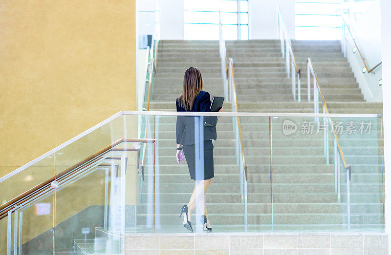 一名身着职业装的匿名女性手持文件夹走上办公楼或公司园区的一段楼梯
