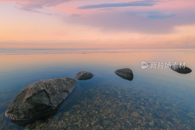 宁静的日落景观在湖岸与粉红色的温暖辉光反射在水，雪和大岩石