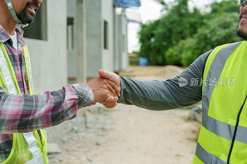 在一个建筑工地，穿着安全背心和头盔的男工人握手，表示同意或完成了一项任务。建筑师和工程师监督房屋建设。