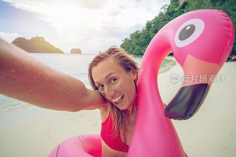 一个年轻的女人在恬静的海滩上与粉红色的火烈鸟在清澈的海水中自拍在泰国群岛。人们旅行奢侈，乐趣和酷的态度概念