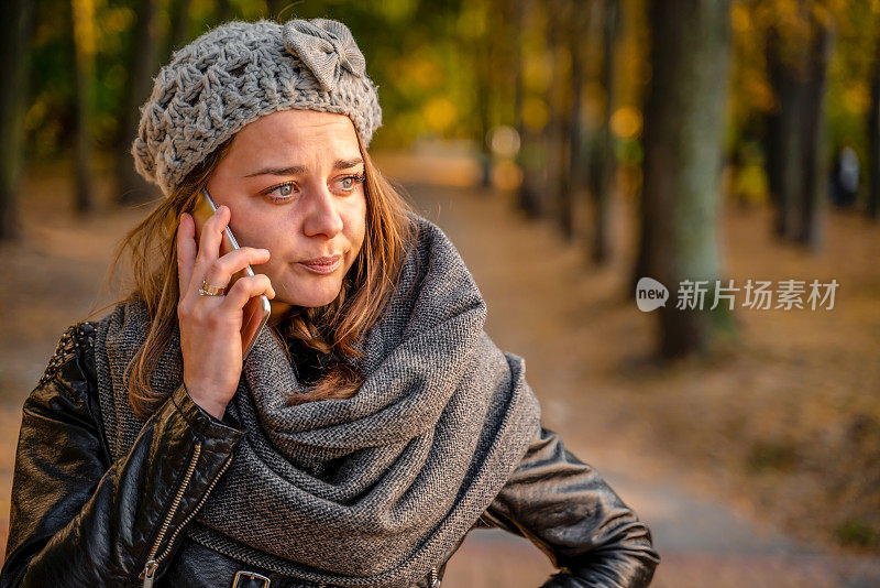 悲伤的女孩站在室外美丽的秋天城市公园里讲智能手机