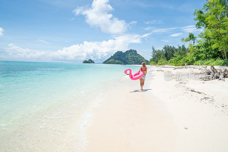 一名年轻女子与充气火烈鸟在泰国群岛田园诗般的海滩上散步。人们旅游目的地有趣和酷的态度概念