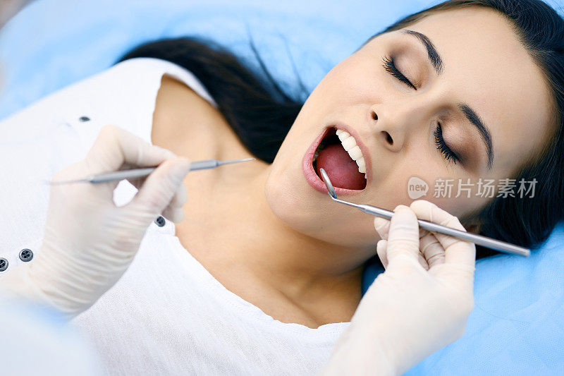 年轻的女病人正在看牙医。美丽的女人与健康的直白牙齿坐在牙科椅子与张大的嘴在口腔检查期间，而医生工作的牙齿