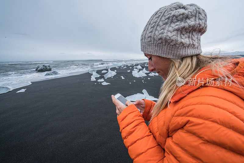 一名年轻女子走在冰岛的黑沙滩上，一边用手机发短信