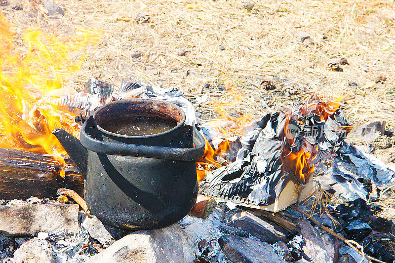 一个黑烟熏过的茶壶在火上，四周是黄色的火舌，背景是干枯的草——徒步旅行的游客库存。