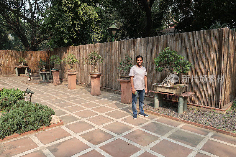 一名英俊的印度男子站在花园中铺成的庭院中，以盆景树为底座展示日本元素
