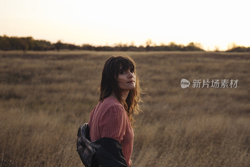 美丽的年轻女子脱下她的皮夹克享受日落在一个高草地