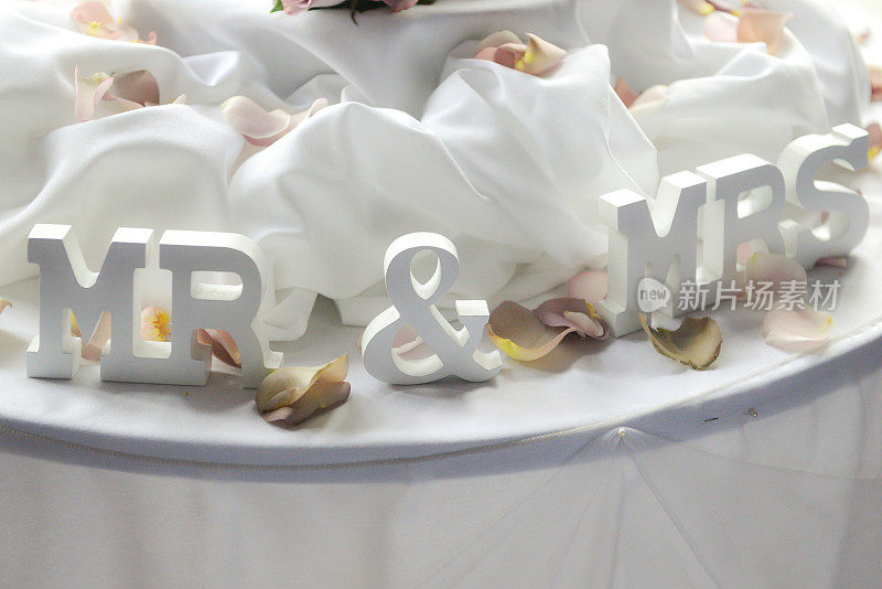 婚礼餐桌上的新婚夫妇点缀着玫瑰花瓣和白色丝绸，作为婚礼蛋糕的美丽背景。