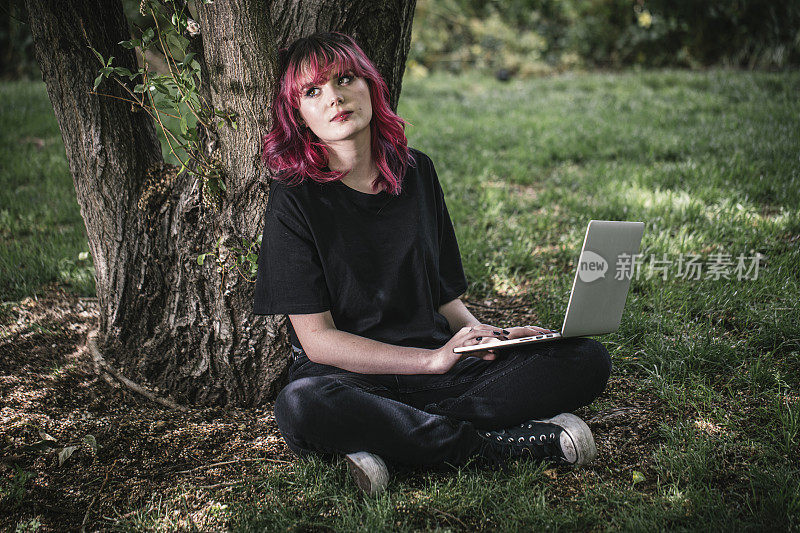 一个年轻的女人用笔记本电脑远程工作，她坐在公园的一棵树下。