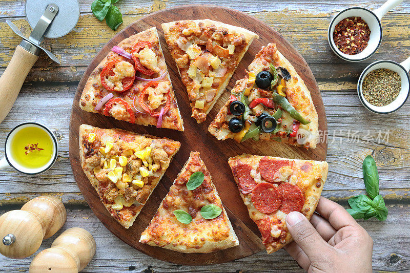 这张图片上的人根本认不出是谁，正在挑选披萨片，各种配料，包括夏威夷披萨、羊乳酪、番茄片和红洋葱、鸡肉和甜玉米、意大利辣香肠、玛格丽塔披萨和圆形木砧板上的黑橄榄蔬菜
