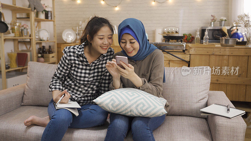 两个多民族朋友坐在家里厨房的客厅沙发上放松。年轻的亚洲中国女孩微笑着，而伊斯兰女子姐姐分享手机屏幕购物网上夏季销售沙发。