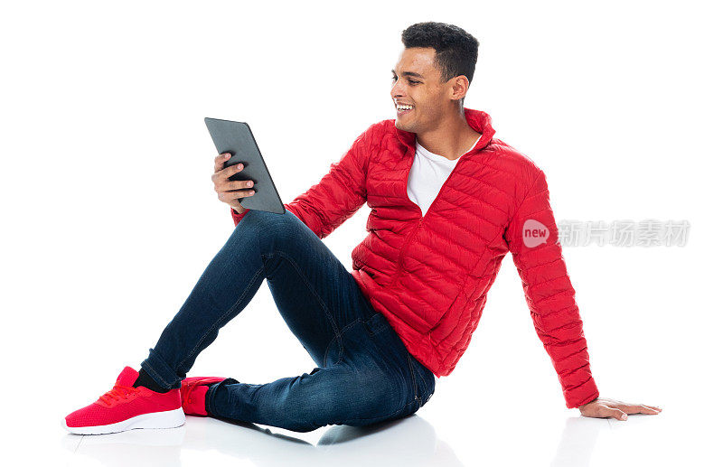 非裔美国青年男性坐在地板上，背景是白色的，穿着牛仔裤，使用数码平板电脑