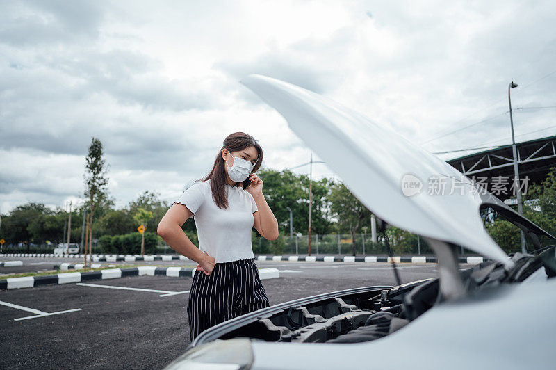 一名年轻的亚洲妇女戴着防护口罩，在她的汽车抛锚前用智能手机寻求帮助