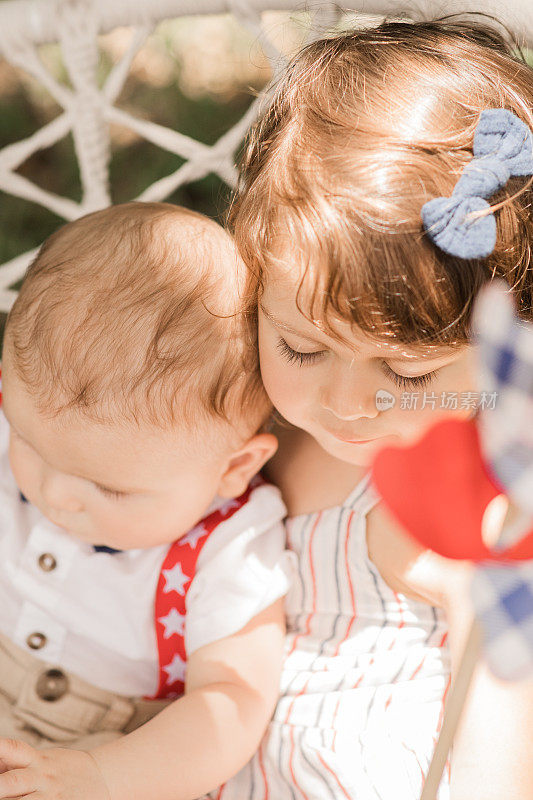 7月4日，一个6个月大的小男孩穿着红色背带和蓝色领结被他3岁的大姐姐抱着和依偎着，她穿着红色，白色和蓝色条纹的裙子，而他们在一个波西奥吊椅上摇摆，玩一个旋转玩具