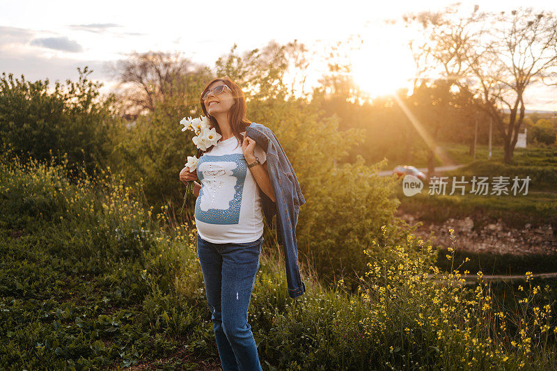 美丽的孕妇手里拿着一束水仙花，在大自然中漫步，欣赏美丽的日落