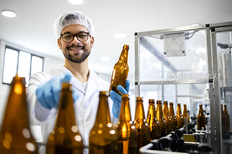 专业的经验丰富的技术工人站在啤酒装瓶厂检查产品质量的画像。在背景生产线上与啤酒瓶机并线。
