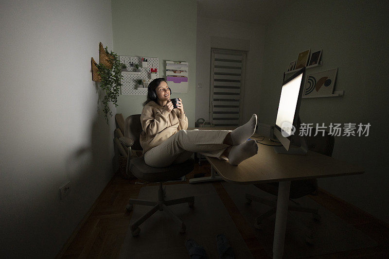 女人坐在家里的办公桌前喝咖啡，用笔记本电脑工作到深夜;在家远程加班的女性网页设计师