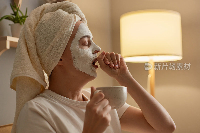 女性在白色毛巾坐在沙发上在家做美容手术，拿着咖啡杯，感到困倦，打哈欠，用手捂住嘴。