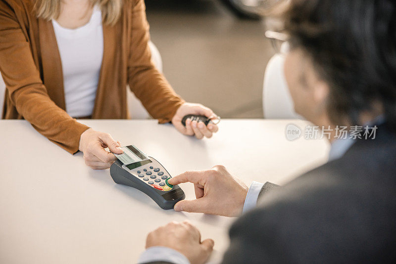 一名女顾客正用信用卡偿还一辆昂贵的汽车，她和一名汽车销售员坐在汽车经销商室内的办公桌旁