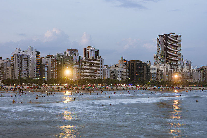 巴西桑托斯市。何塞·梅尼诺海滩。下午晚些时候，海滨建筑和拥挤的海滩和冲浪者在水中冲浪。长曝光摄影。