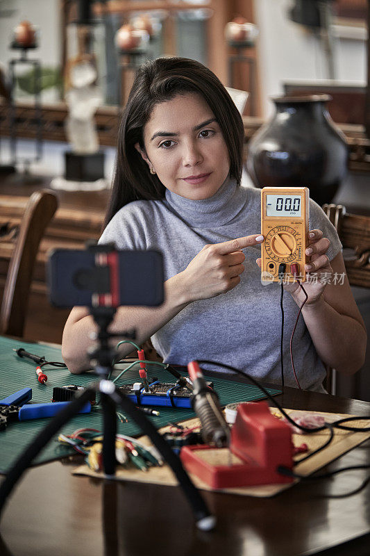 西班牙裔年轻女子在家教电子维修的特写。Youtuber展示了在使用智能手机进行在线培训时使用万用表。