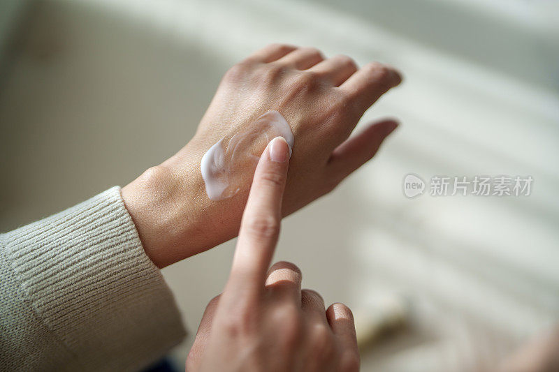 在这张剪短的图片中，一位中年亚洲女性正在用润肤霜涂抹双手干燥的皮肤，阳光温柔地照在她的皮肤上，突显出滋养的姿态。