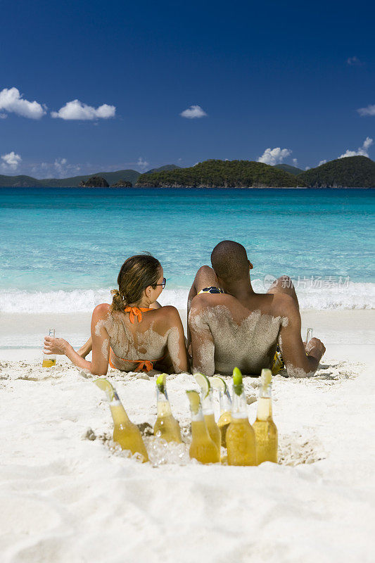 一对情侣在热带海滩上喝着啤酒，晒着日光浴