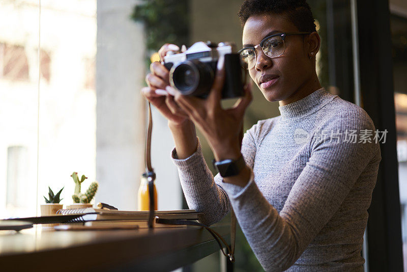 集中的非洲裔美国女性摄影师拿着复古相机拍摄现代设计的咖啡馆室内工作坐在靠近窗户的木桌上笔记本和文具