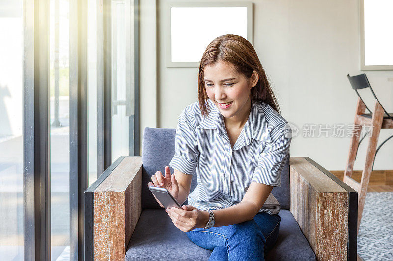快乐的亚洲女商人坐在工作空间，使用智能手机在现代的办公室或咖啡店。企业技术的概念