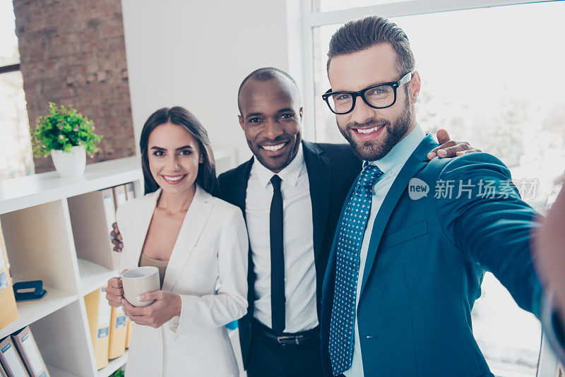 三个伙伴的自拍照，胡子男人在眼镜前自拍与他的同事在经典的西装，拿着杯子和茶在手里，站在工作场所，站
