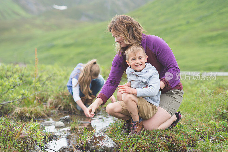 一位妈妈和她的孩子们在他们的徒步旅行中休息一下，去看看小路旁边的小溪。