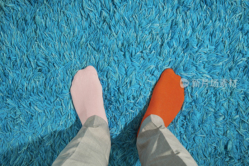 POV的观点，男人的脚穿着奇怪的袜子，粉色和橙色，对着一个明亮的蓝色地毯。