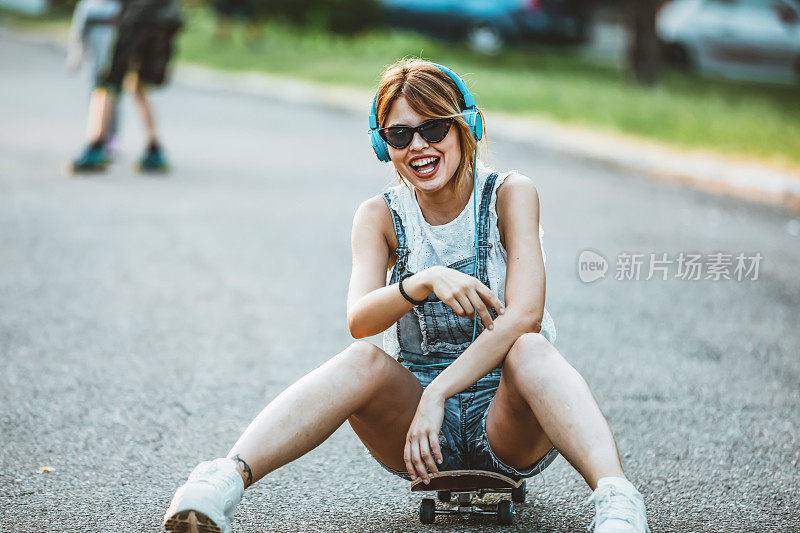 年轻女子戴着耳机坐在路上的滑板上。在一个阳光明媚的日子里，在公园里玩滑板的漂亮年轻女子。年轻迷人的美丽和时尚的年轻女人与滑板摆姿势的照片。她站在C里享受