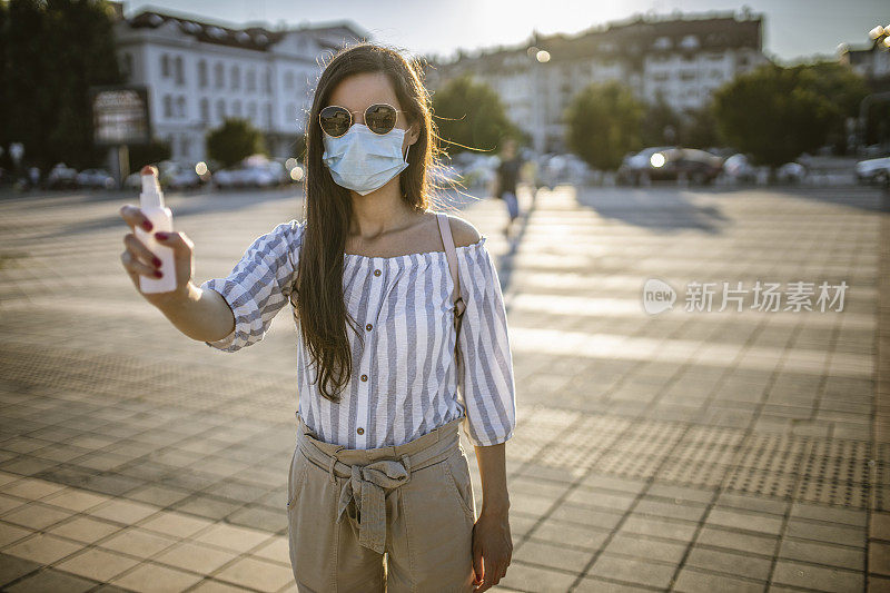 一名年轻女子戴着外科口罩，手上涂着洗手液，在封锁期间站在空荡荡的城镇广场上