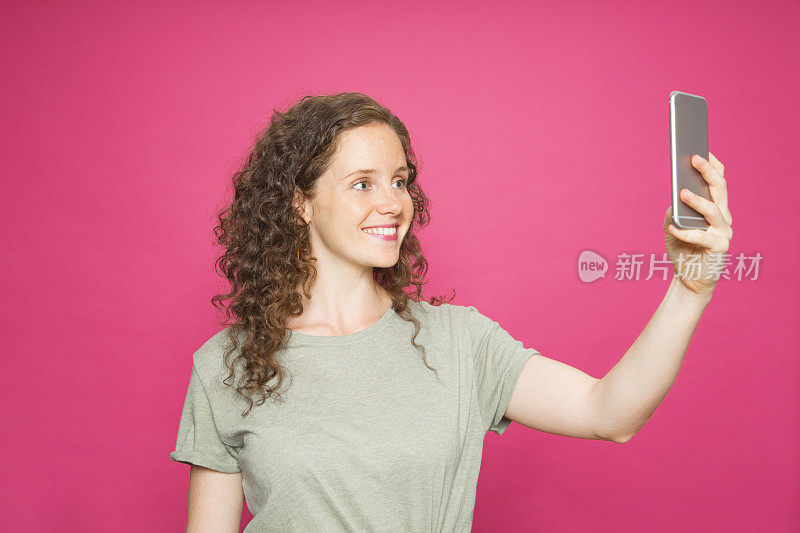 一名年轻女子手拿手机，粉红色背景上穿着绿色t恤，拍了一张自画像