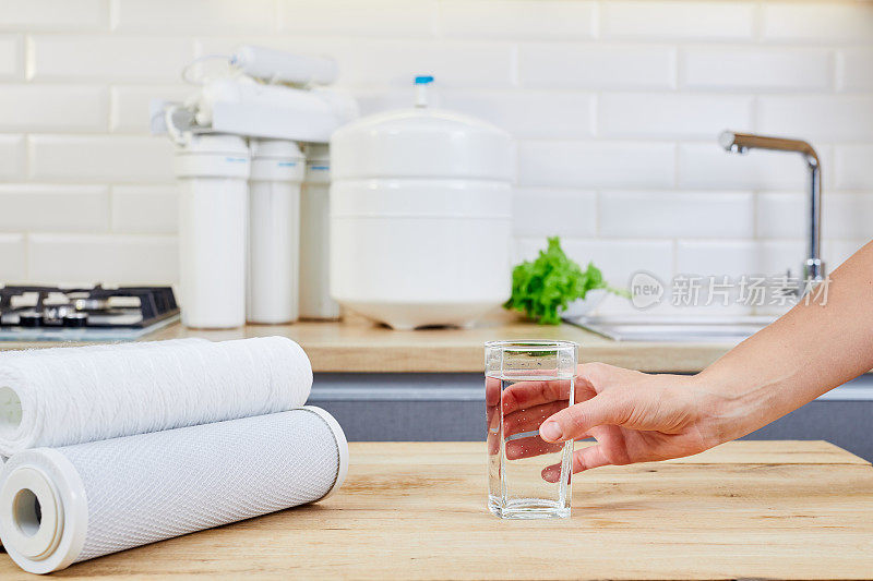在厨房的餐桌上，拿着一杯纯净水的女人。女性的手握着透明的水杯。背景水过滤器。Hydratation。