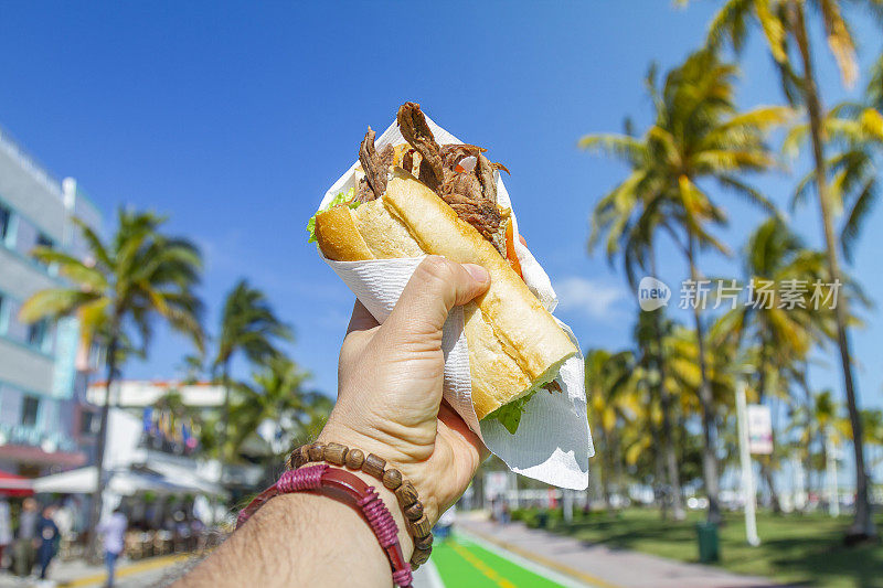 一个年轻的旅行男性享受他的假期，吃着古巴三明治在海洋大道前，南海滩，迈阿密，迈阿密，南佛罗里达，美国。