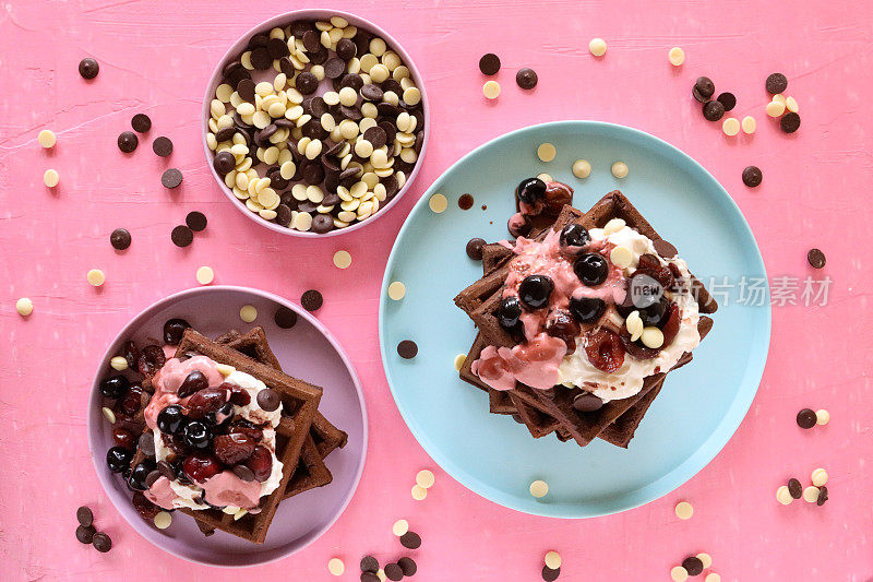 一堆堆自制的巧克力华夫饼，上面放着融化的覆盆子冰淇淋、蓝莓和樱桃，牛奶和白巧克力片，淡紫色和浅蓝色的盘子，粉红色的背景，令人赏心悦目