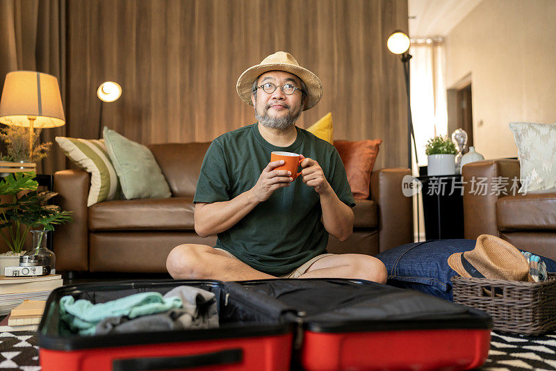 亚洲胡子男成年人戴着帽子享受打包布放在行李箱里的行李准备新的旅行假期带着微笑开朗在家里的客厅里，亚洲男人手拿热咖啡放松休闲收拾完行李箱