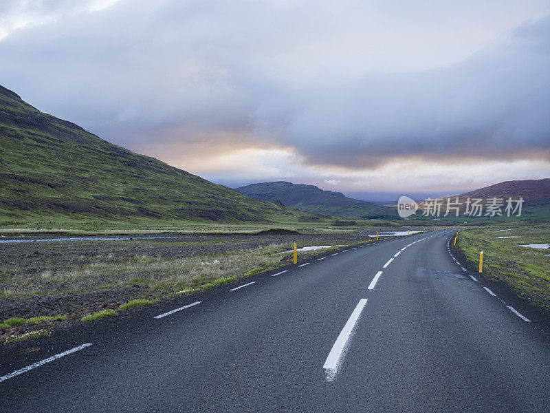 柏油路弯道穿越空旷的北部景观与绿草如茵的五彩山丘、水坑和夕阳映衬的天空，一路延伸至冰岛西部的山地高地，复制空间