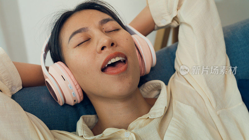 特写年轻的亚洲女子闭着眼睛躺在沙发上休息戴着耳机听音乐唱着最喜欢的歌在音频流媒体广播，在家在线播客。