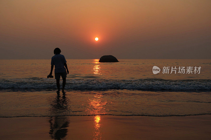 印度果阿的Palolem，印度果阿，度假游客赤脚走在海滩上，手里拿着鞋子在水里飞溅，橙色日落，后视图，重点在前景，复制空间